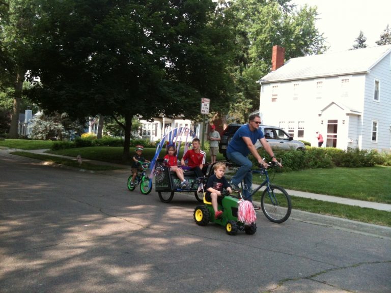 Boy driving a toy tractor alongside Dad pulling a cart with a seat, decorated for ECCO 4th of July Parade 2015
