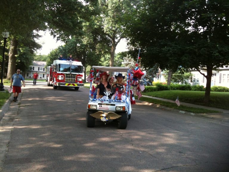 Parade participants in golf cart decorated with flags, and red, white, and blue bunting and balloons