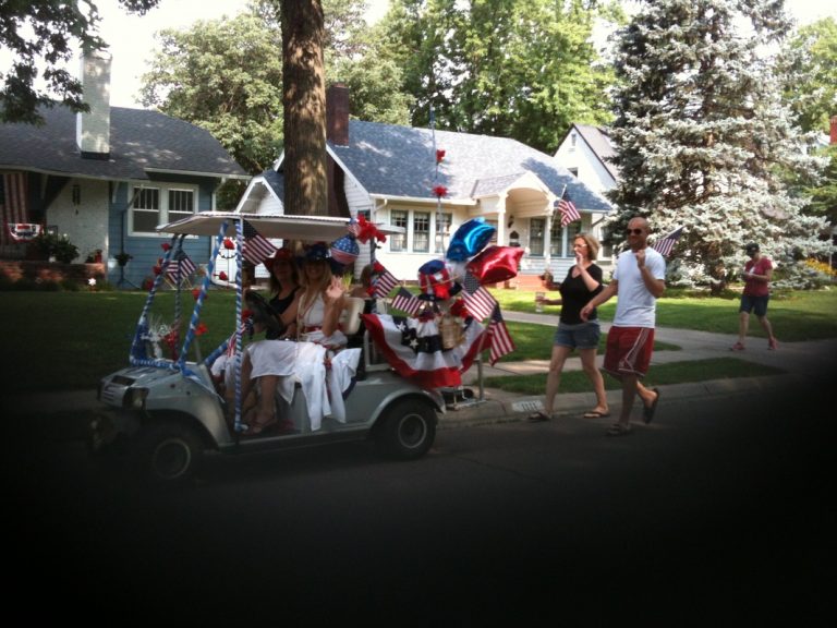 Parade participants in golf cart decorated with flags, and red, white, and blue bunting and balloons